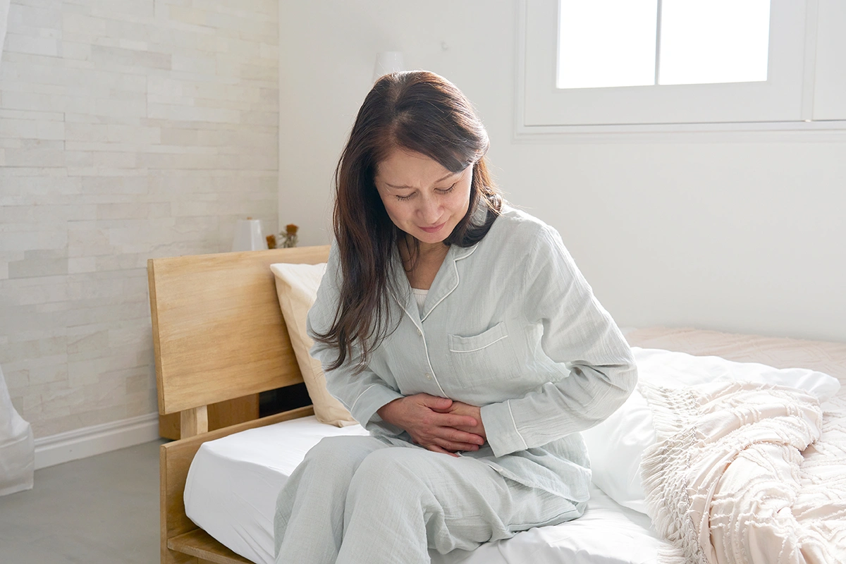 Asian woman in pajamas sitting and holding her stomach with a pained expression.